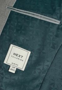 Chaqueta de traje ajustada en tejido verde azulado, con un patrón geométrico, acentos grises, y una etiqueta con la información de talla y marca.