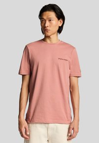Camiseta de algodón de color rosa claro con mangas cortas, cuello redondo y texto negro "LYLE & SCOTT" en el pecho. Textura suave, diseño casual.