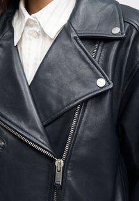 Κοντινό πλάνο μαύρου δερμάτινου μπουφάν με ασημένιο φερμουάρ και κουμπιά, που φοριέται πάνω  από λευκό πουκάμισο με λεπτές μπλε ρίγες.