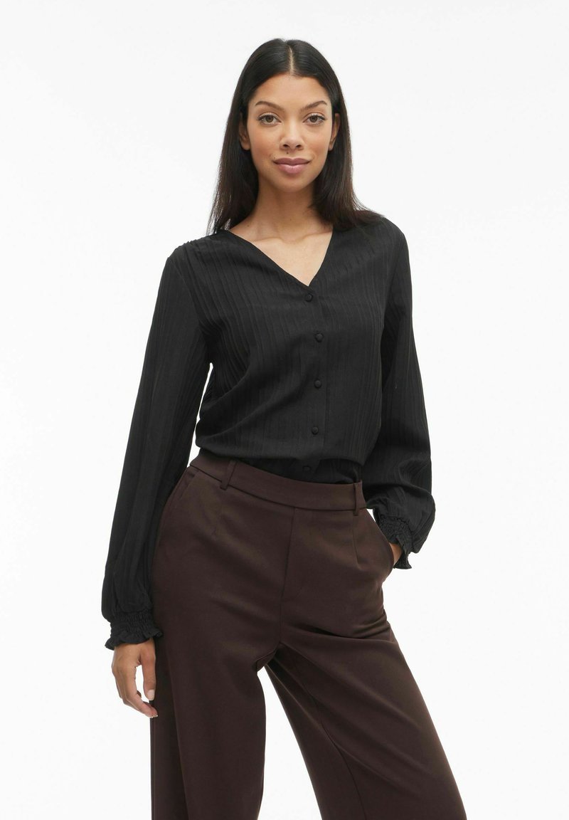 Melnas blūzes ar garām piedurknēm ar vertikālām svītrām, V veida izgriezumu, priekšpuses pogām un pūslīšveida aprocēm, kopā ar brūnām biksēm ar augstu jostasvietu.