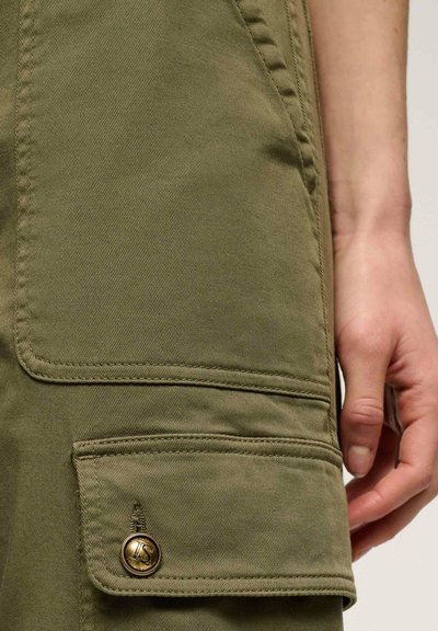 Dettagli dei pantaloni cargo verde oliva con una grande tasca a toppa e hardware in bronzo. Il materiale è cotone testurizzato con cuciture visibili.