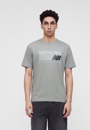 Jeune homme aux cheveux bouclés portant un t-shirt gris New Balance Athletics et un pantalon foncé sur un fond clair uni.