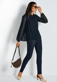 Navybluse mit transparenten Ärmeln, kombiniert mit figurbetonten navyfarbigen Hosen. Hält eine braune Tasche mit dekorativen runden Akzenten. Beigefarbene spitze Pumps.
