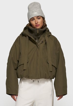Olivgrüne, verkürzte Jacke mit einem Kordelzug in der Kapuze, die zwei vordere Taschen und einen Reißverschluss am Hals hat. Graue Strickmütze. Weiße Hose.