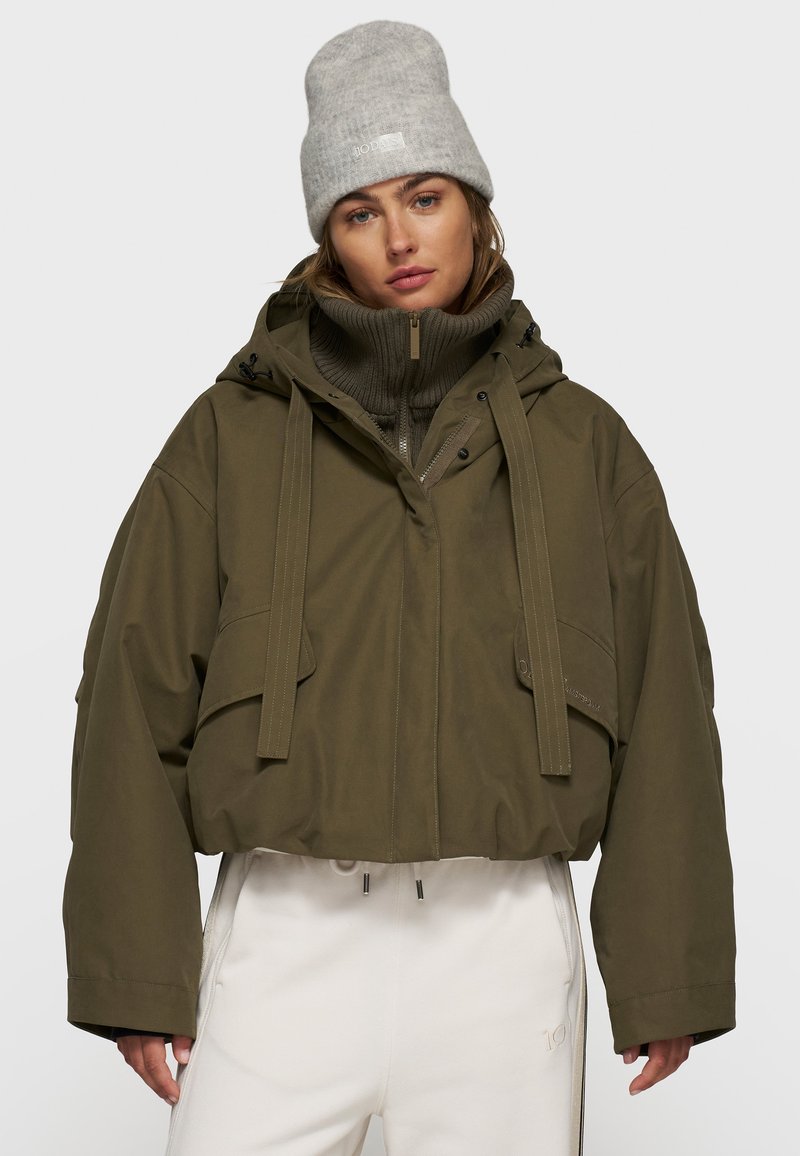 Veste cropped vert olive avec capuche à cordon de serrage, dotée de deux poches avant et d'un col zippé. Bonnet en tricot gris. Pantalon blanc.