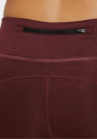 Mörkröda leggings med en slät yta, som har en liten ficka med dragkedja på baksidan och kontrasterande sömmar längs kanterna.