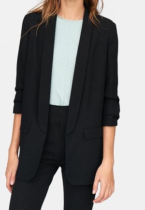 Frau trägt schwarzen Blazer mit hochgekrempelten Ärmeln über hellblauem gemustertem Oberteil und schwarze Hose, steht mit entspannt an den Seiten hängenden Händen.