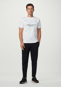 Bílý tričko s potiskem "HACKETT SPORT" a geometrickým vzorem, doplněné černými teplákovými kalhotami a černými sportovními botami.