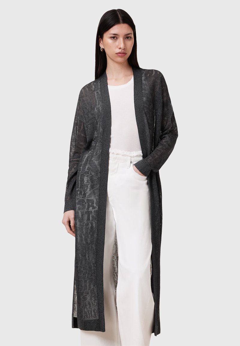 Lång, öppen cardigan framtill i mörkgrått, med ett texturerat mönster, långa ärmar och sidoflikar, bärs över en ljus topp och krämfärgade byxor.