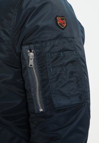 Manche de veste marine avec poche zippée, compartiments cousus, et un patch rouge et or comportant des lettres stylisées sur le bras supérieur.