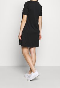 Donna che indossa un corto vestito nero e sneaker bianche, in piedi con le gambe incrociate, vista da dietro su uno sfondo semplice.