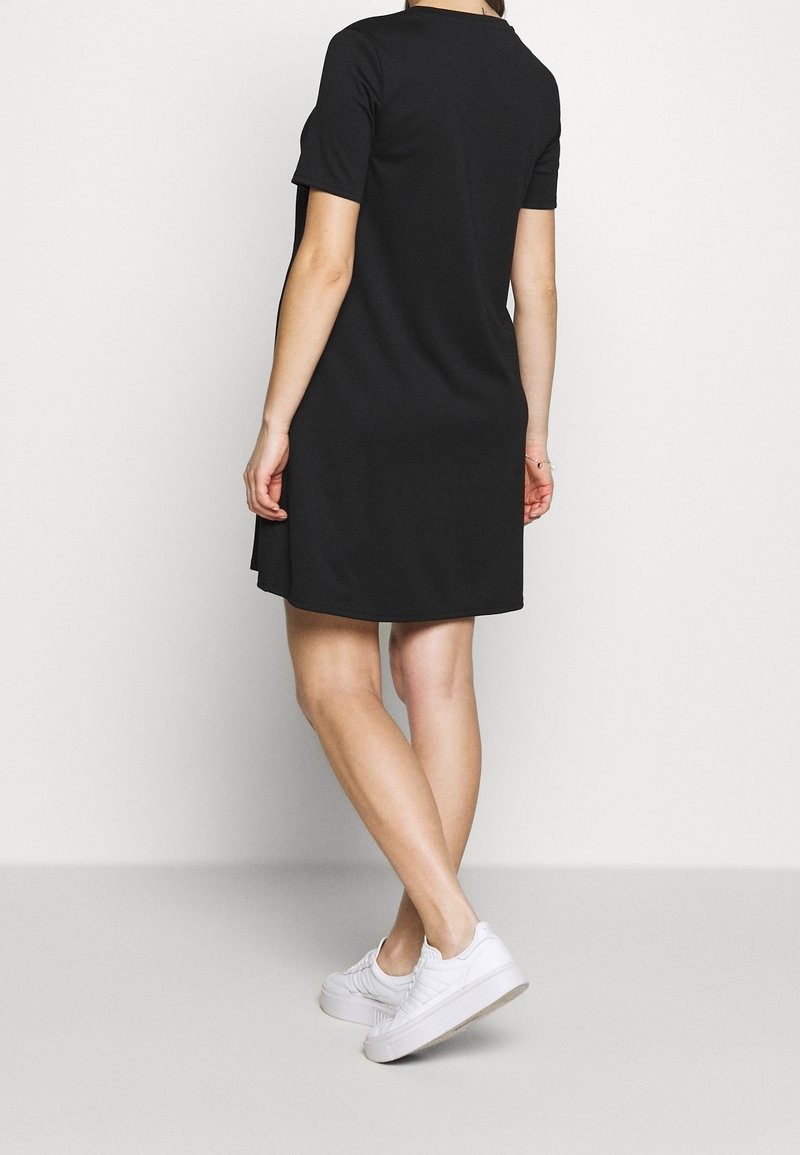 Donna che indossa un corto vestito nero e sneaker bianche, in piedi con le gambe incrociate, vista da dietro su uno sfondo semplice.