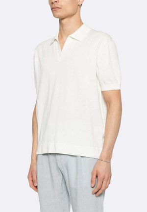 Uomo che indossa una polo bianca a maniche corte e pantaloni grigio chiaro, in piedi davanti a uno sfondo chiaro e uniforme.