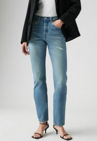 Jean taille haute bleu clair à coupe slim, avec des détails déchirés et un ourlet effiloché, associés à des talons noirs à lanières.