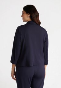 Granatowa marynarka o miękkim materiale, z wyraźnym kołnierzem, rękawami o długości trzech czwartych oraz zawiniętymi mankietami. Posiada czystą konstrukcję szwu tylniego.