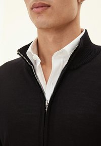 Black zip-up sweater with a ribbed collar over a white shirt. The sweater features a smooth texture and silver zipper detailing.