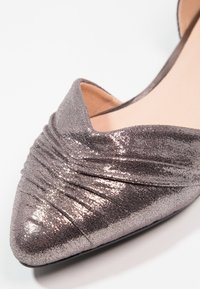 Chaussure plate gris métallisé à bout pointu, avec une texture froncée et un matériau lisse et brillant. Les côtés ouverts rehaussent le design.