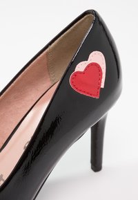 Chaussure à talon haut en cuir verni noir avec des patchs superposés en forme de cœur rouges et roses sur le côté extérieur près du talon.