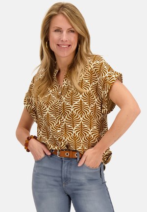 Vrouw glimlacht met lang blond haar, draagt een bruin-wit patroon kortemouwenblouse, blauwe jeans en een barnstenen armband, handen in de zakken.