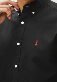 Chemise noire à boutons en coton avec des boutons blancs et un petit logo de homard rouge sur la poitrine gauche. Elle présente un design de col classique.