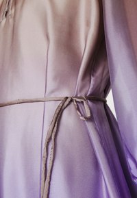 Robe dégradée lavande avec une texture lisse, des manches longues et une fermeture à nouer. Le tissu a un léger éclat et une drapé doux.