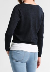 Suéter de punto navy con escote barco, con costura texturada en los hombros, llevado sobre una camisa blanca, combinado con jeans de color azul claro.