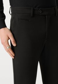 Czarne spodnie wykonane z gładkiej tkaniny mają płaski przód, boczną kieszeń oraz pojedyncze zapięcie na guzik. Dłoń lekko spoczywa na udzie.