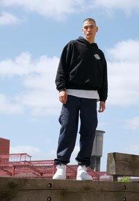 Svart hettegenser med logo, hvit t-skjorte under, marineblå cargo-bukser og hvite sneakers, som står ute mot en blå himmel.