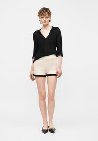 Femme portant un haut noir à manches trois-quarts, un short crème texturé avec bordure noire, des lunettes et des ballerines noires pointues, debout devant un fond blanc.