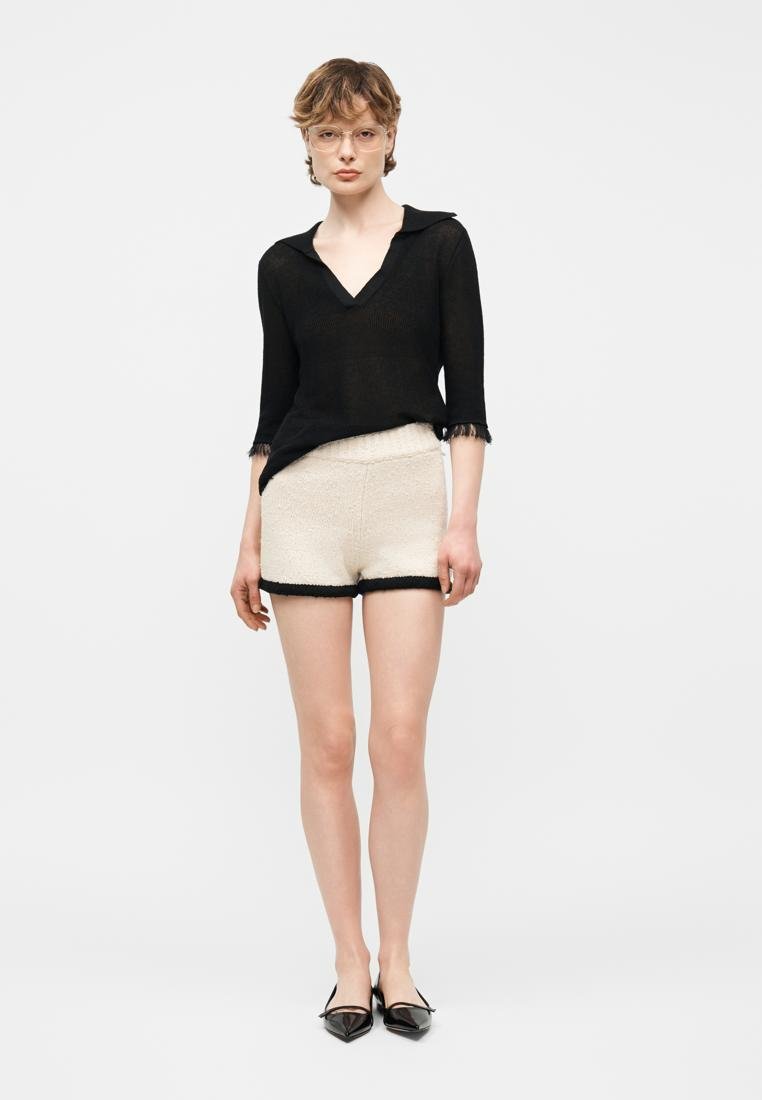 Femme portant un haut noir à manches trois-quarts, un short crème texturé avec bordure noire, des lunettes et des ballerines noires pointues, debout devant un fond blanc.