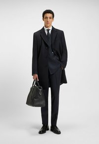 Cappotto di lana navy con design doppiopetto, completo su misura, camicia bianca, cravatta navy e una borsa in pelle nera; scarpe eleganti nere lucide.