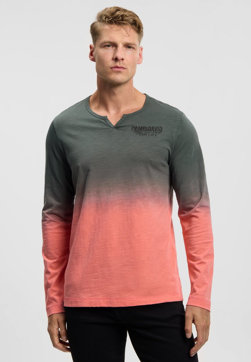 Young man with short curly hair wearing a long-sleeve gradient shirt fading from dark gray to coral, standing against a plain background.