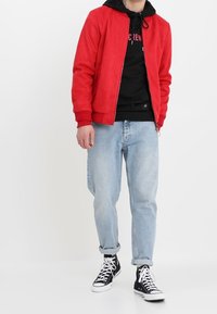 Rode Suède bomberjack met geribde manchetten, gedragen over een zwarte hoodie. Gecombineerd met lichtblauwe jeans en zwarte hoge sneakers.