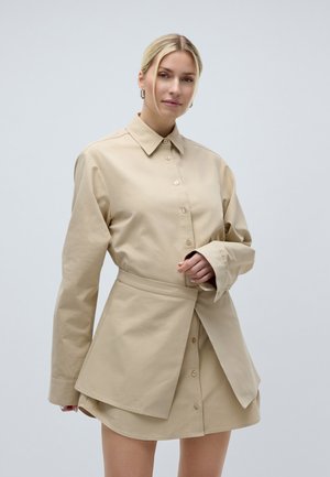 Woman with blonde hair wearing a beige oversized button-up shirt dress with a flared layered skirt detail and hoop earrings.