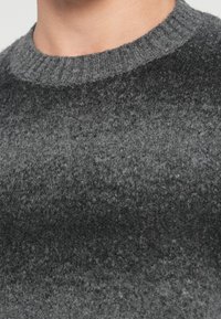 Γκρι, υφασμένο πουλόβερ από μάλλινη ξηρή που διαθέτει στρογγυλή λαιμόκοψη με ραβδώσεις. Διαθέτει μια μαλακή, αφράτη επιφάνεια με διάφορες αποχρώσεις του γκρι.