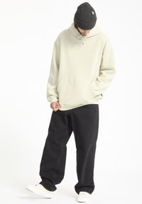 Lichtgroene hoodie met een voorzak, trekkoord en klein logo. Gecombineerd met losse zwarte broek en witte sneakers. Geweven beanie.