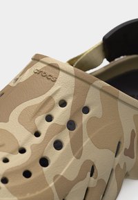 Κοντινή λήψη της κάμελου και μπεζ καμουφλάζ Crocs clog με οπές αερισμού και ρυθμιζόμενο λουράκι στη φτέρνα.
