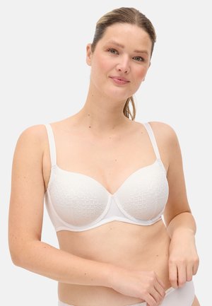 Mujer de piel clara y cabello castaño, vestida con un sujetador de encaje blanco, de pie contra un fondo claro y liso, ajustándose la ropa interior.