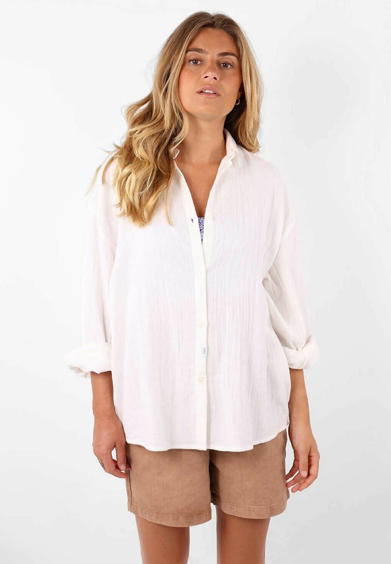 Chemise blanche à manches retroussées, en tissu texturé et avec une coupe décontractée, assortie à un short tan. Aucun motif ou accent visible.