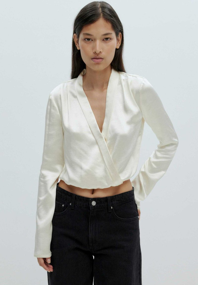 Crème-kleurige satijnen wikkeltop met lange mouwen, diepe V-halslijn en cropped lengte, gecombineerd met hoge taille zwarte denim jeans.