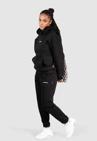 Schwarzer Hoodie mit Fronttasche und Logo; kombiniert mit passenden Jogginghosen mit seitlichen Reißverschlusstaschen und Branding. Weiße Sneaker.