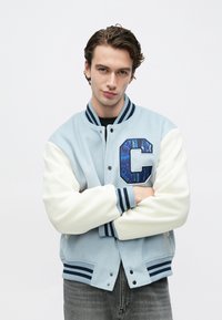 Νέος άνδρας με σταυρωμένα τα χέρια φοράει ανοιχτό μπλε varsity μπουφάν με λευκά μανίκια και μεγάλο διακοσμητικό γράμμα «C» στο στήθος.