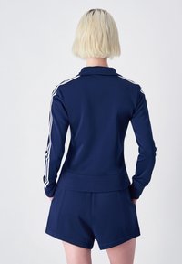 Giacca sportiva blu con strisce bianche lungo le maniche, polsini e colletto a coste, abbinata a pantaloni corti coordinati con tasche laterali.