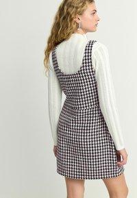 Melna un rozā houndstooth kleita ar pieguļošu dizainu, platām siksnām un noapaļotu muguru. Valkāta virs strukturēta balta garām piedurknēm tops.