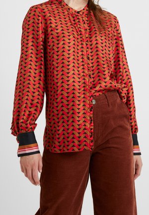 Frau trägt eine langärmelige Bluse mit roten und orangefarbenen geometrischen Mustern und braune Cordhosen.