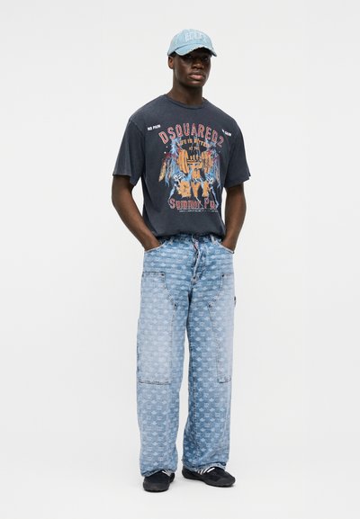 Άνδρας που φορά μπλε καπέλο μπέιζμπολ, μαύρο μπλουζάκι με γραφικά, ανοιχτό μπλε τζιν παντελόνι με πλατιά πόδια και μαύρα αθλητικά παπούτσια, στέκεται με τα χέρια στις τσέπες.