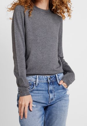 Person med krøllet hår, der bærer en grå langærmet sweater og blå slidte jeans med den ene hånd i lommen og den anden hvilende nedad.