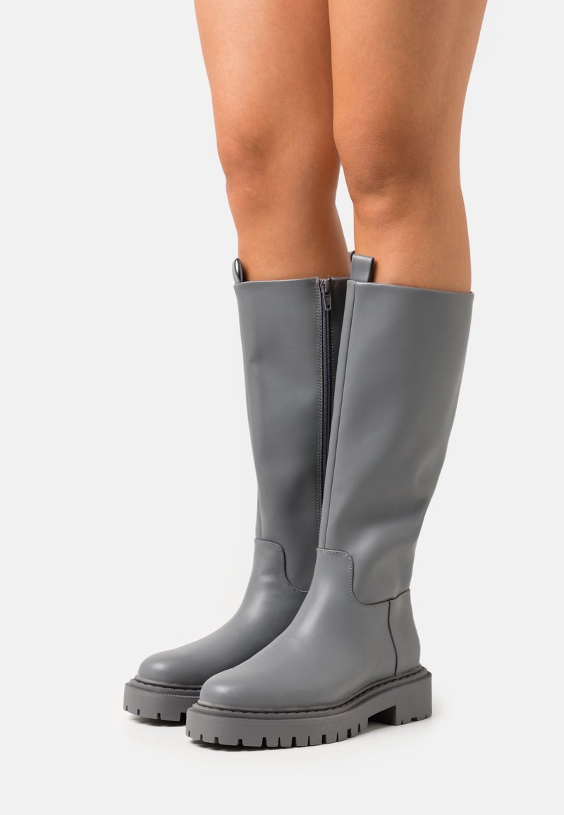 Jambes portant des bottes en cuir gris hautes jusqu'aux genoux avec des fermetures éclair latérales et des semelles épaisses à crampons, debout sur un fond clair uni.