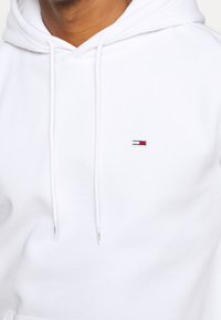Sudadera blanca hecha de tela suave, con un parche de logo en tonos rojo y azul. Incluye cordones y un diseño de corte relajado.