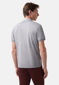 Šedé polo tričko s krátkým rukávem, s jednoduchým límečkem a hladkou texturou. Nosené s tmavě hnědými kalhotami, zobrazující pohodlný pohled zezadu.